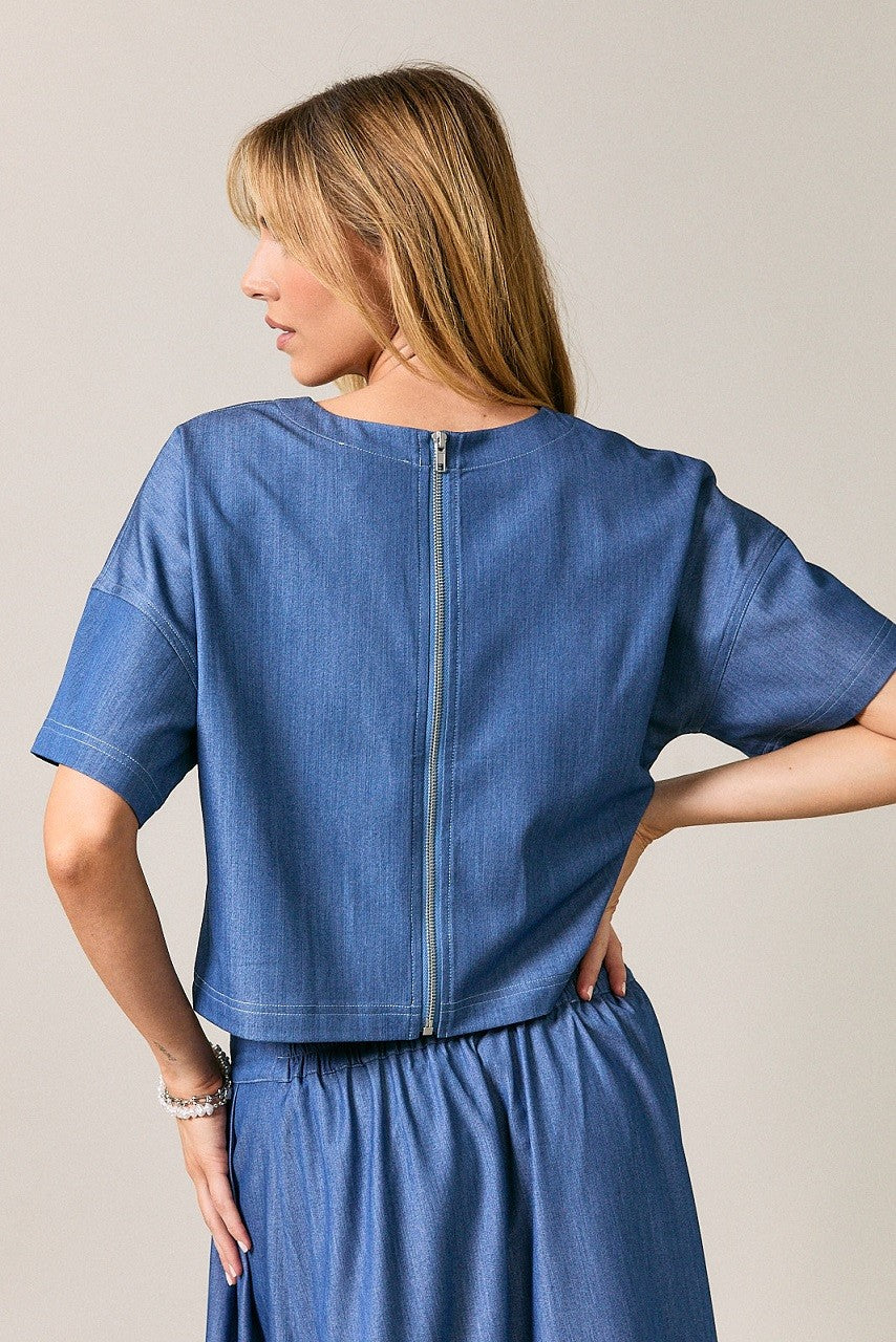 Back view of a woman wearing a cropped short-sleeve top with back zipper against a plain light gray studio background.