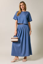 A woman wearing a cropped short-sleeve top and matching pleated skirt against a plain light gray studio background.