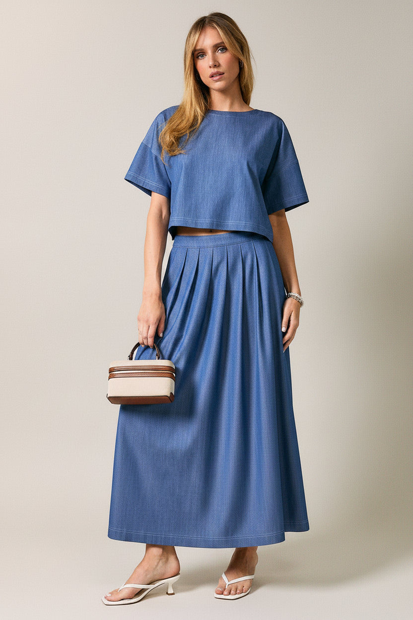 A woman wearing a cropped short-sleeve top and matching pleated skirt against a plain light gray studio background.