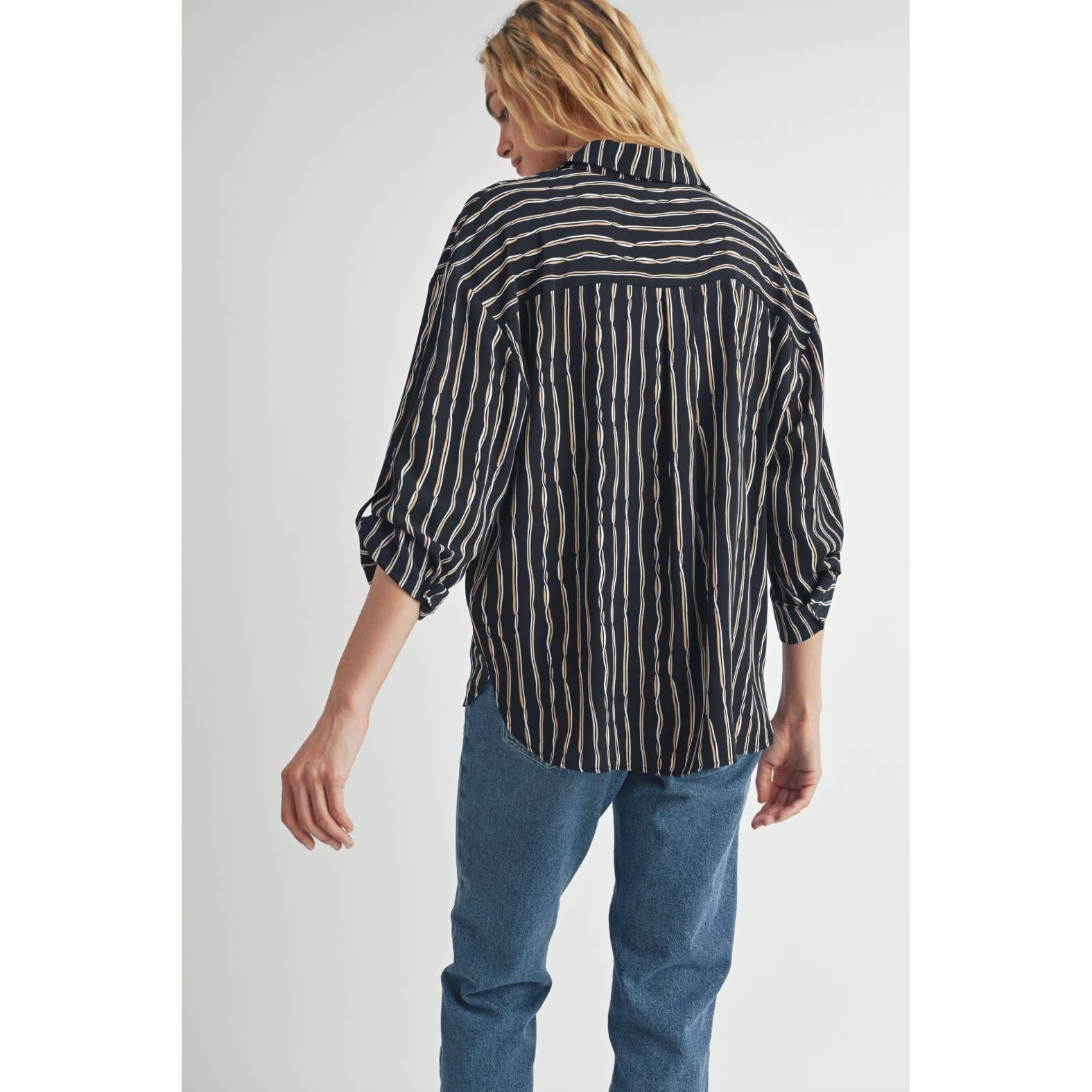 Woman wearing a black blouse with ivory and tan stripes and blue jeans on a plain background