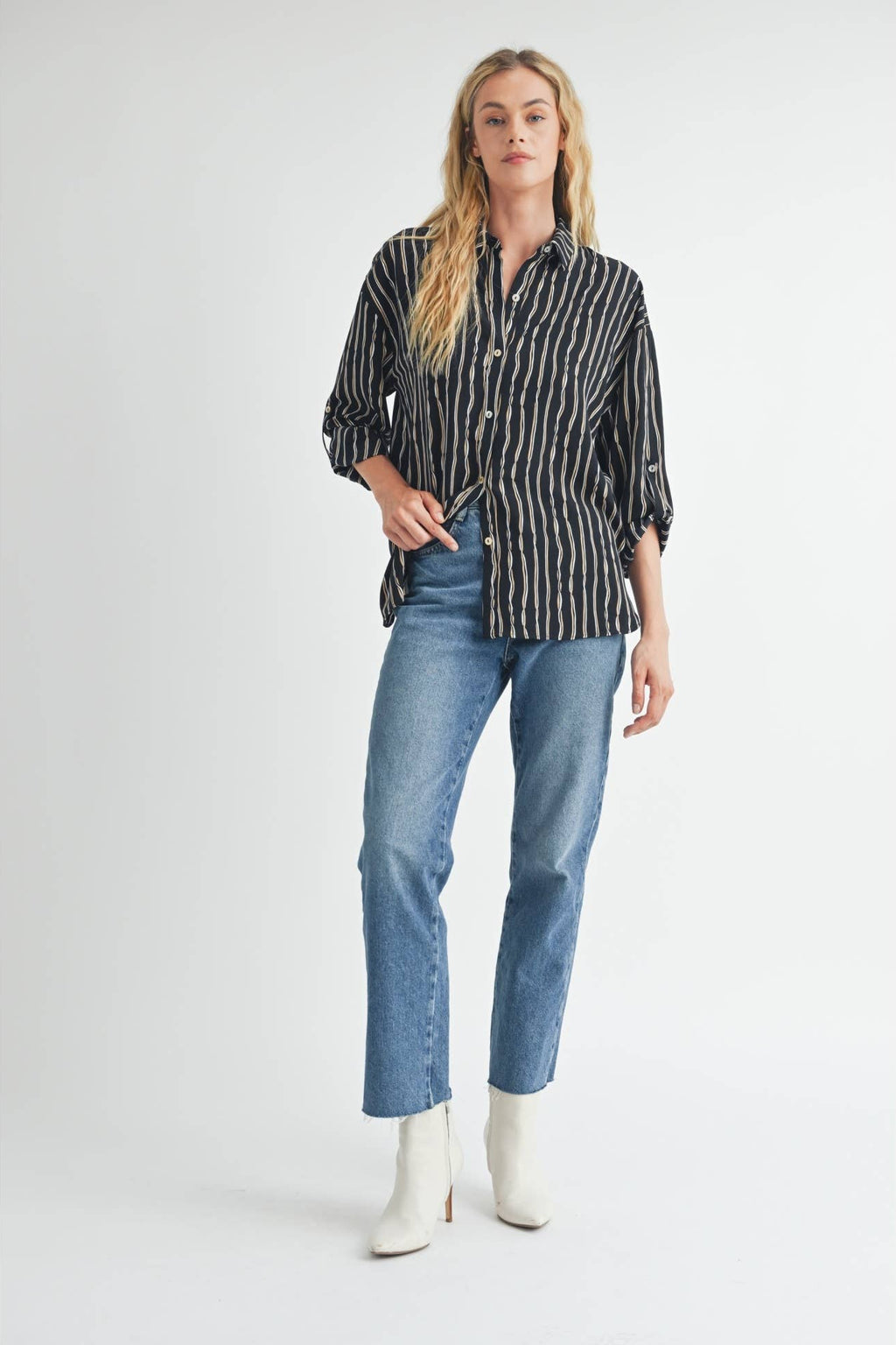 Woman wearing ivory and tan stripes on black blouse, jeans, and white boots on a plain background