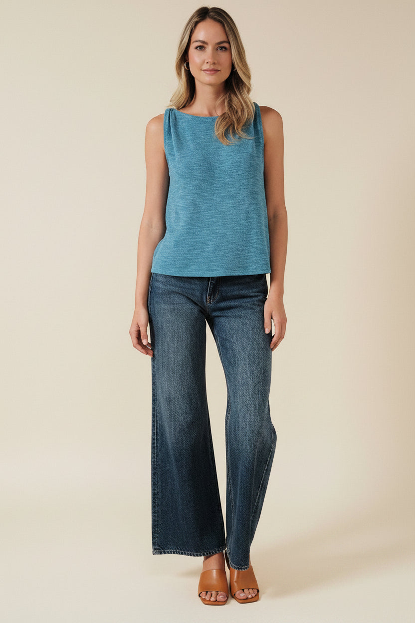 Woman wearing a blue turquoise sleeveless top and dark blue jeans against a beige background