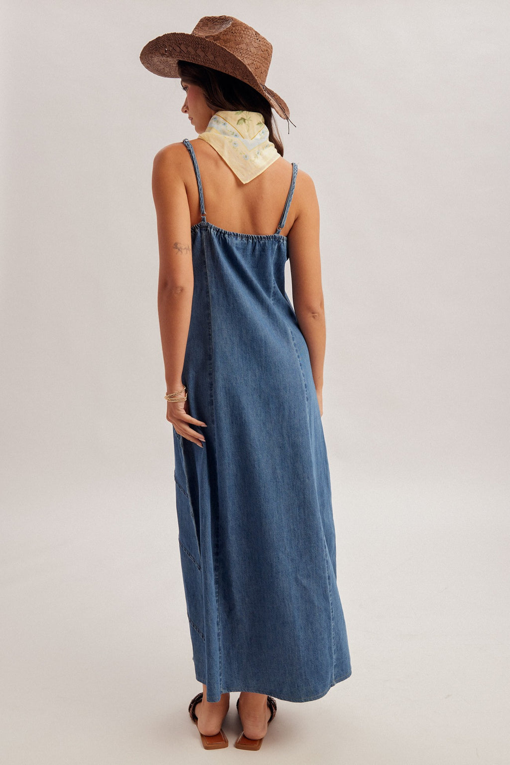 Woman wearing a medium blue denim maxi dress with pintuck detailing and brown hat on a plain background