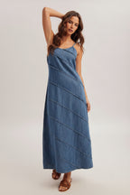 Woman wearing a medium blue denim maxi dress with pintuck detailing and brown hat on a plain background