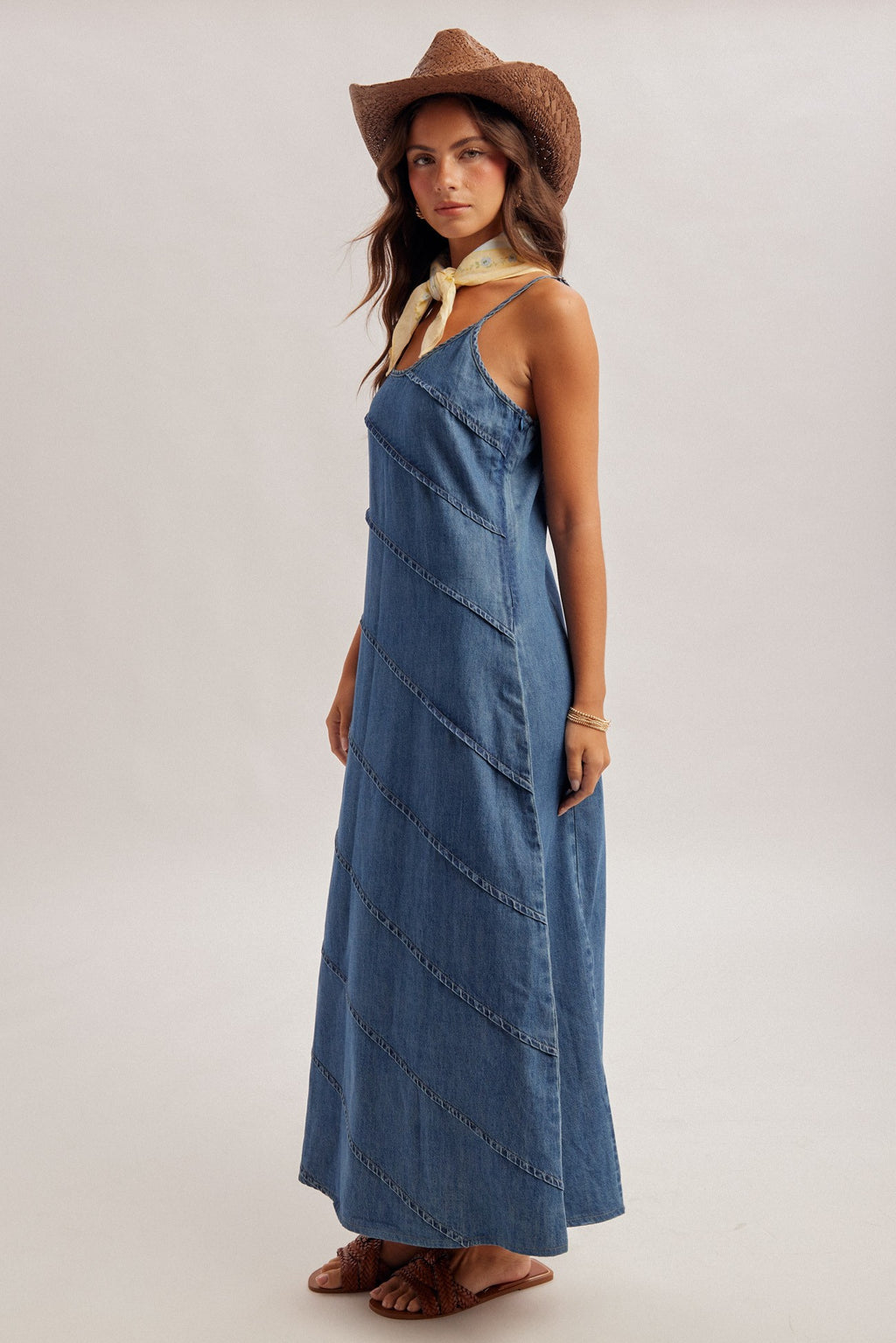 Woman wearing a medium blue denim maxi dress with pintuck detailing and brown hat on a plain background
