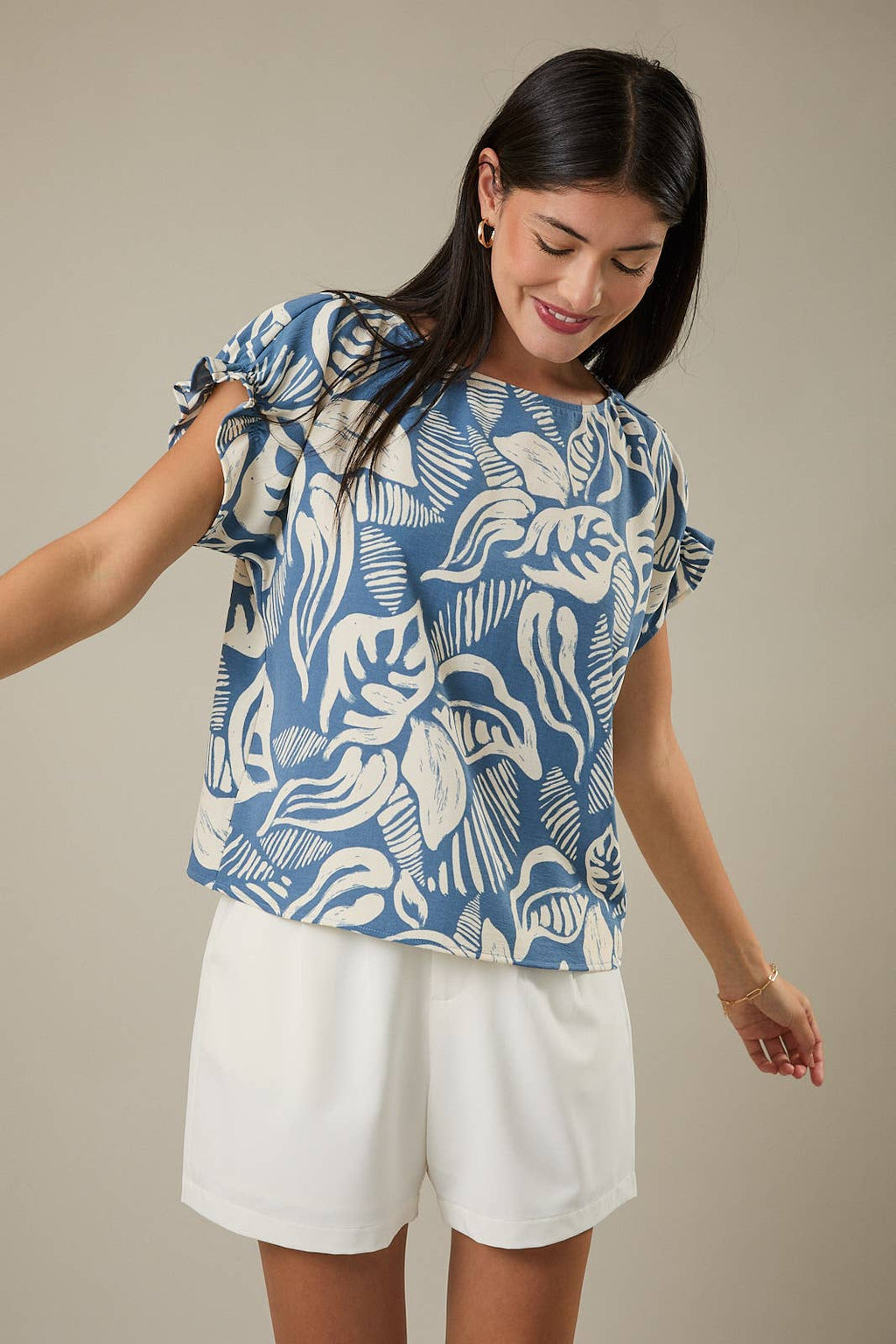 Woman wearing a denim blue and ivory leaf design top and white shorts against a beige background