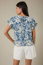 Woman wearing a denim blue and ivory leaf design top and white shorts against a beige background