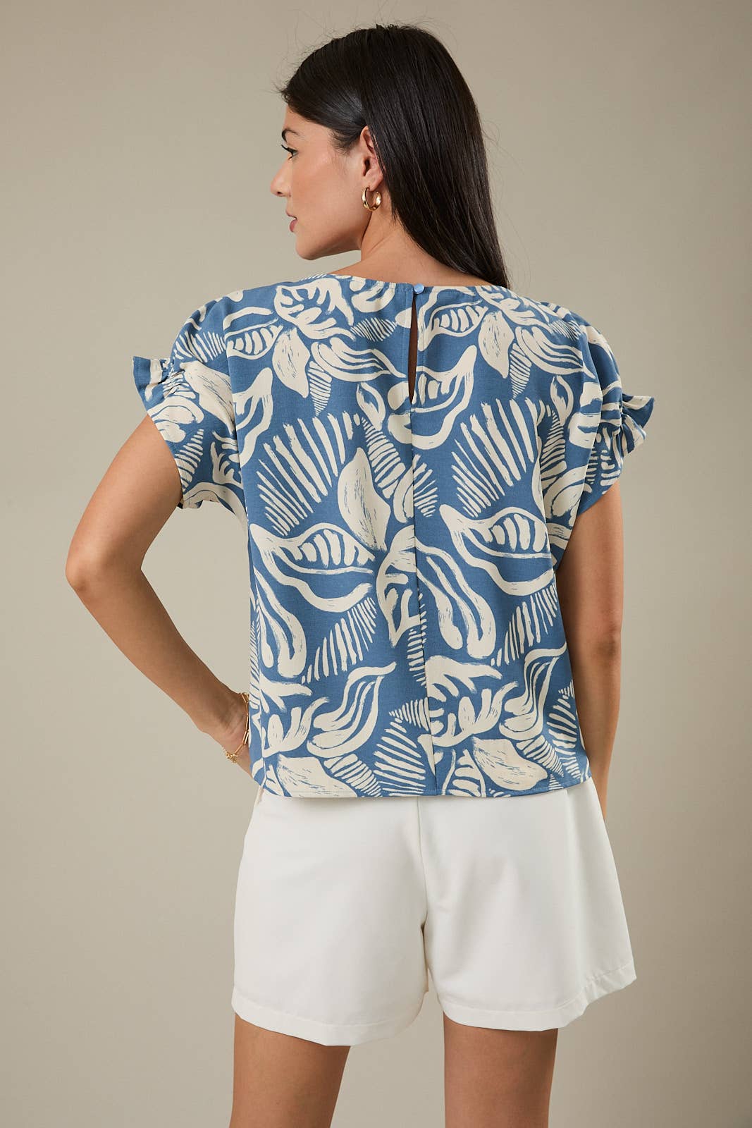 Woman wearing a denim blue and ivory leaf design top and white shorts against a beige background