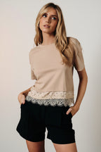 A woman modeling a taupe short-sleeve top with a lace hem detail, paired with black shorts, posed in front of a draped brown fabric backdrop.