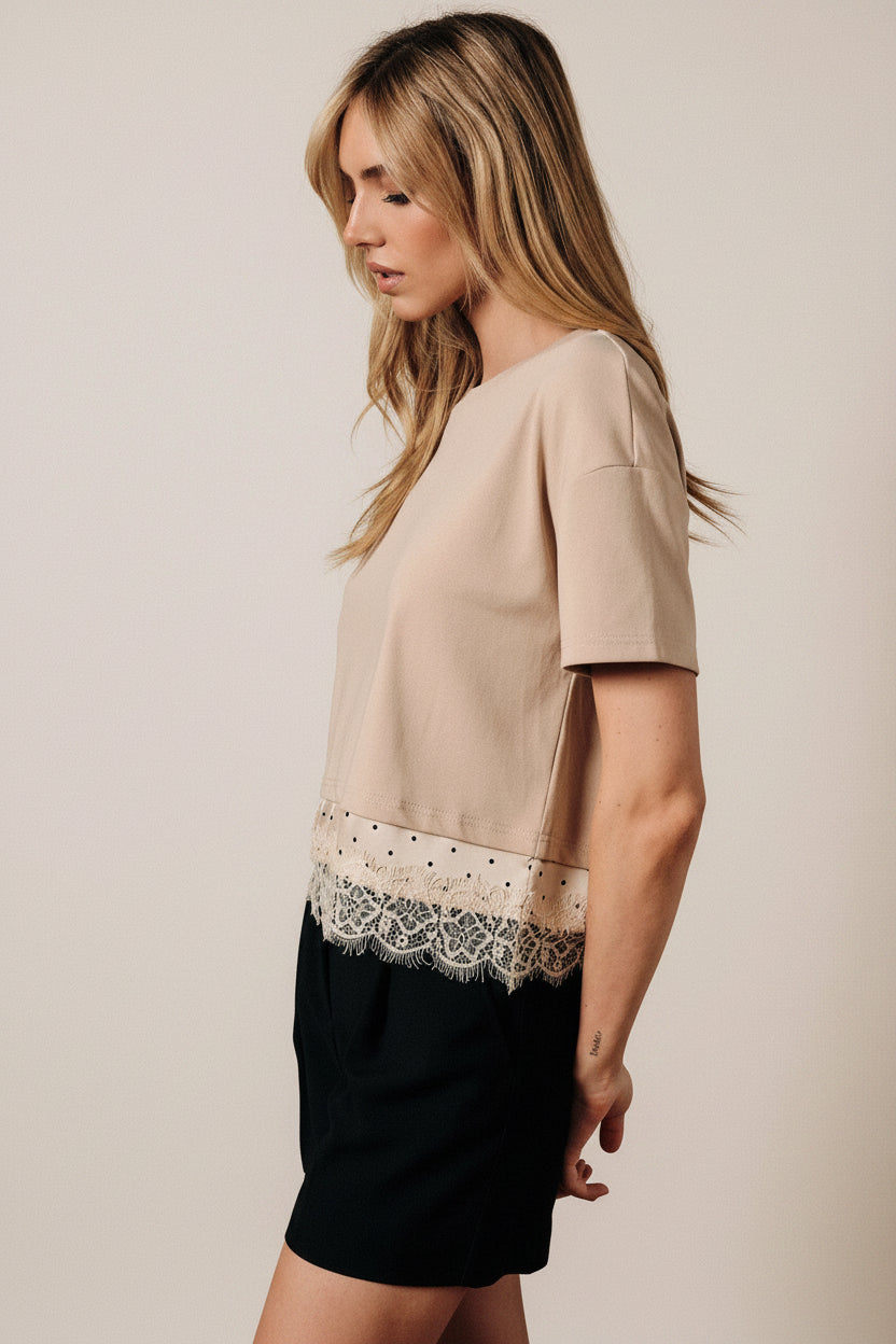 A woman modeling a taupe short-sleeve top with a lace hem detail, paired with black shorts, posed in front of a draped brown fabric backdrop.