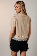 Back view of a woman modeling a taupe short-sleeve top with a lace hem detail, paired with black shorts, posed in front of a draped brown fabric backdrop.