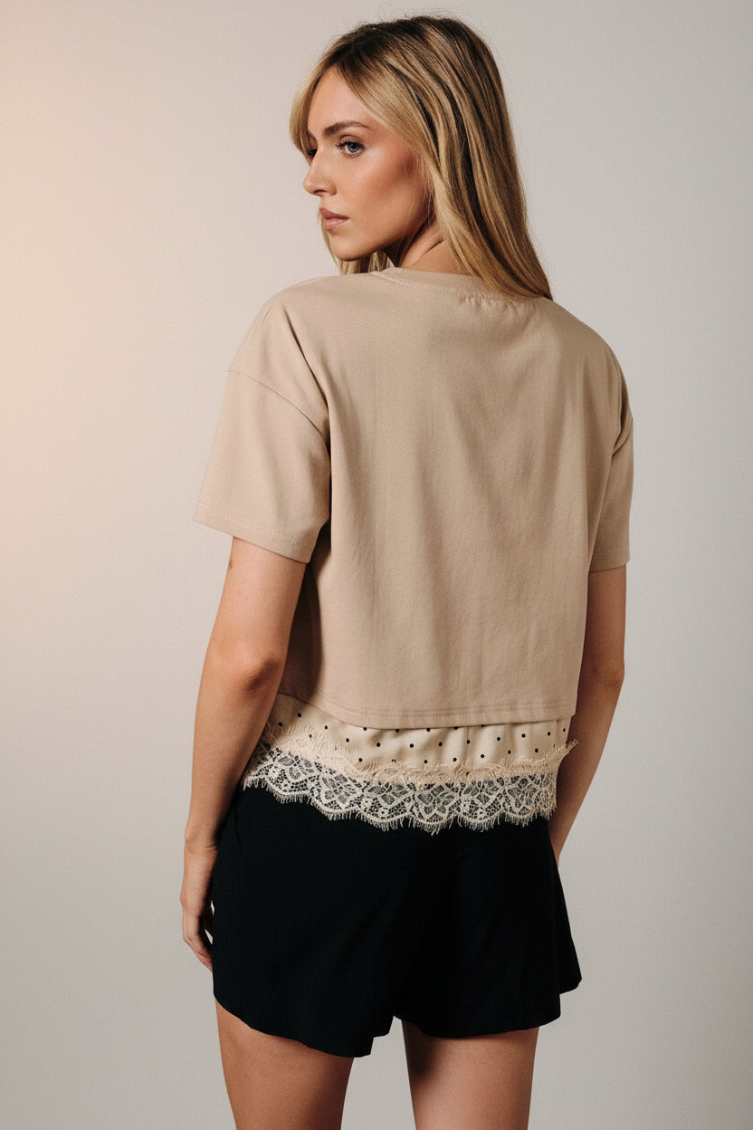 Back view of a woman modeling a taupe short-sleeve top with a lace hem detail, paired with black shorts, posed in front of a draped brown fabric backdrop.