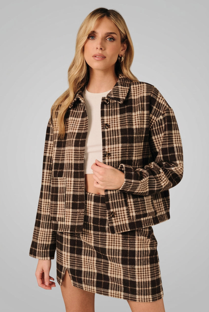 A woman modeling a brown and cream plaid button-up shirt jacket with a matching mini skirt over a white crop top, set against a solid black background.