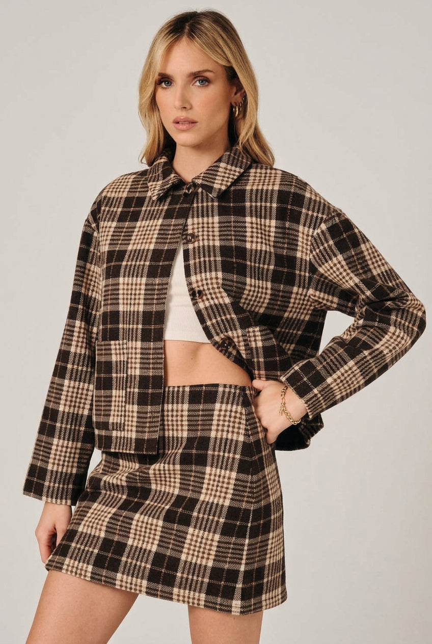 A woman modeling a brown and cream plaid button-up shirt jacket with a matching mini skirt over a white crop top, set against a solid black background.