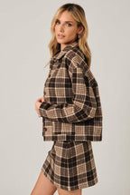 A woman modeling a brown and cream plaid button-up shirt jacket with a matching mini skirt over a white crop top, set against a solid black background.