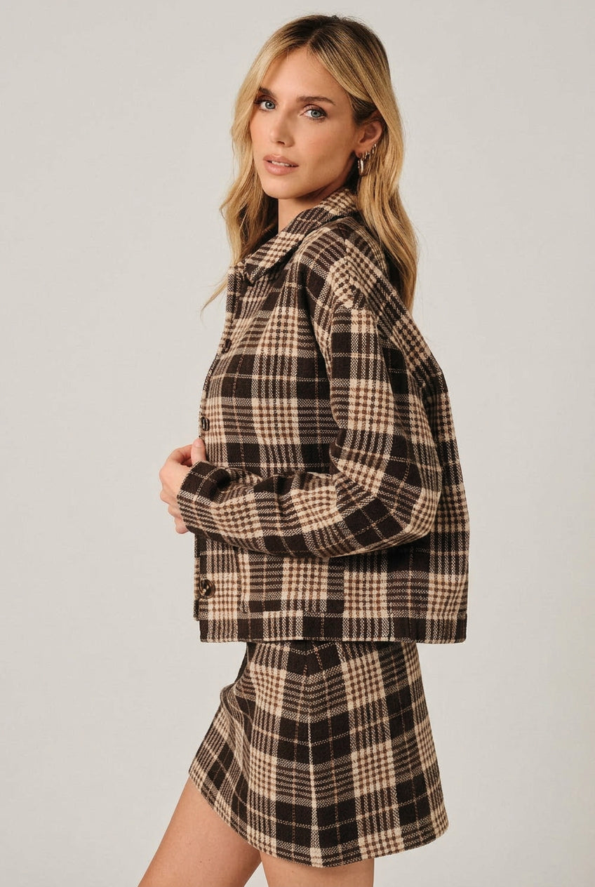 A woman modeling a brown and cream plaid button-up shirt jacket with a matching mini skirt over a white crop top, set against a solid black background.