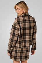 Back view of a woman modeling a brown and cream plaid button-up shirt jacket with a matching mini skirt over a white crop top, set against a solid black background.