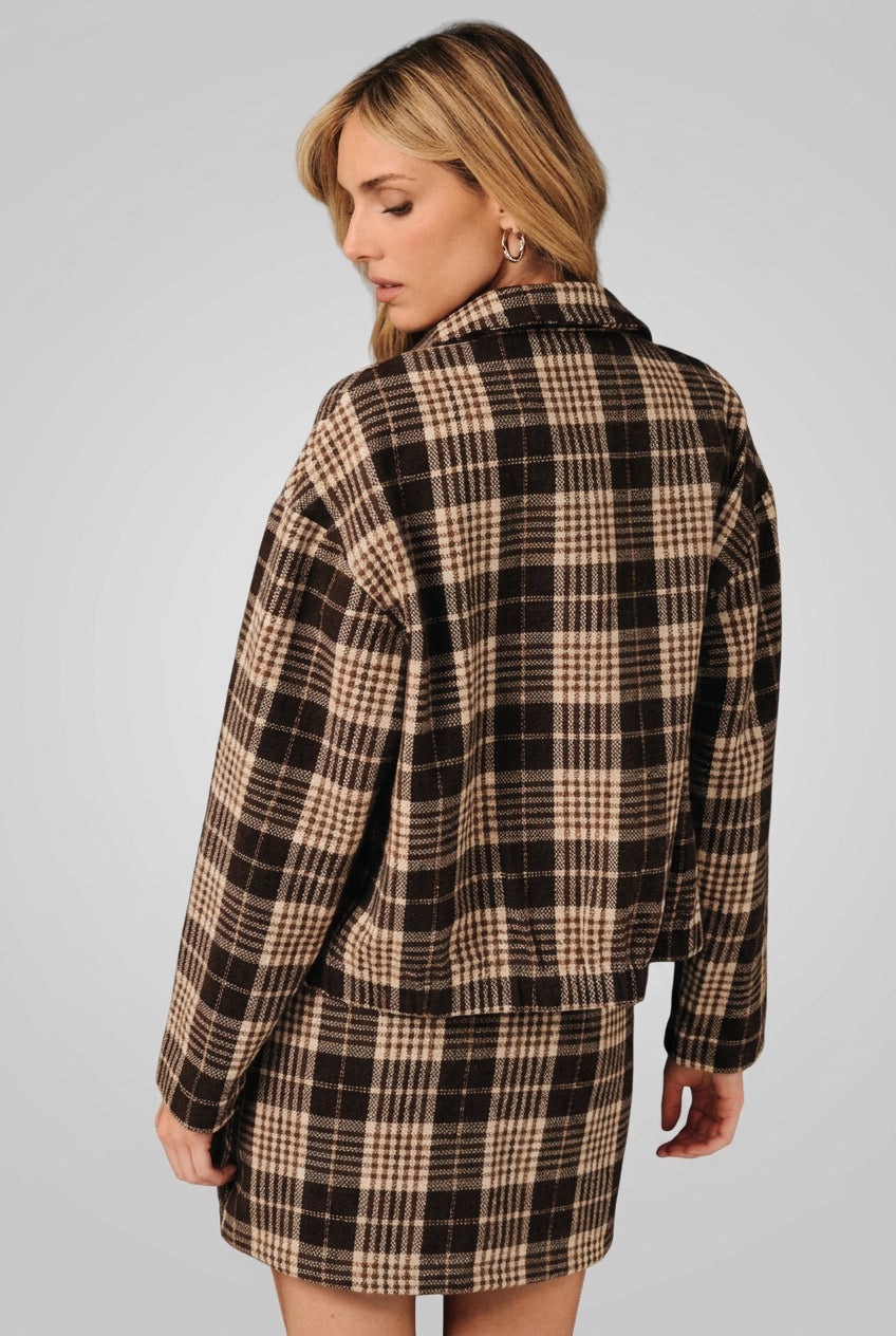 Back view of a woman modeling a brown and cream plaid button-up shirt jacket with a matching mini skirt over a white crop top, set against a solid black background.