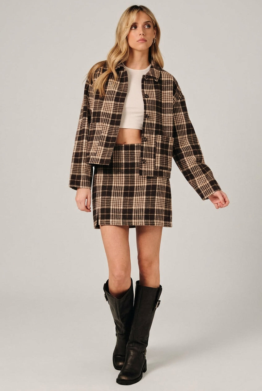 A woman modeling a brown and cream plaid button-up shirt jacket with a matching mini skirt over a white crop top, set against a solid black background.