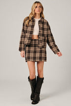 A woman modeling a brown and cream plaid button-up shirt jacket with a matching mini skirt over a white crop top, set against a solid black background.