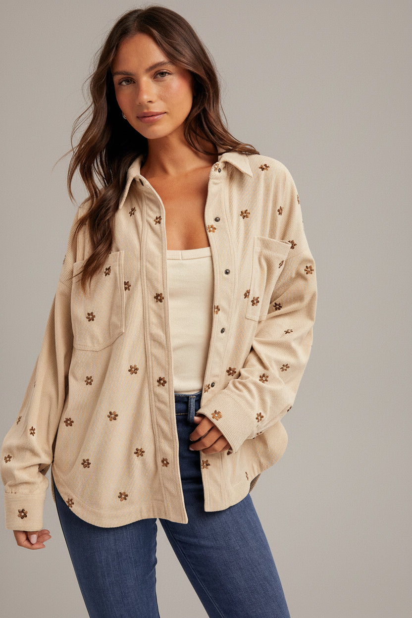 Woman wearing an ecru shirt jacket with brown metallic embroidery floral patterns, brown hat, and blue jeans on a plain background