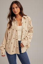 Woman wearing an ecru shirt jacket with brown metallic embroidery floral patterns, brown hat, and blue jeans on a plain background