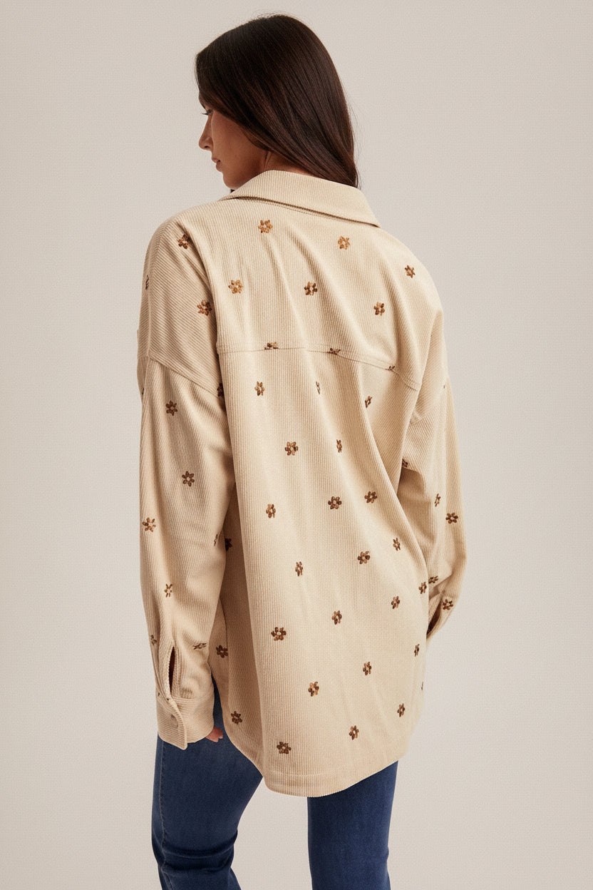 Back of a woman wearing an ecru shirt jacket with brown metallic embroidery floral patterns, brown hat, and blue jeans on a plain background