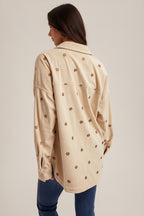 Back of a woman wearing an ecru shirt jacket with brown metallic embroidery floral patterns, brown hat, and blue jeans on a plain background