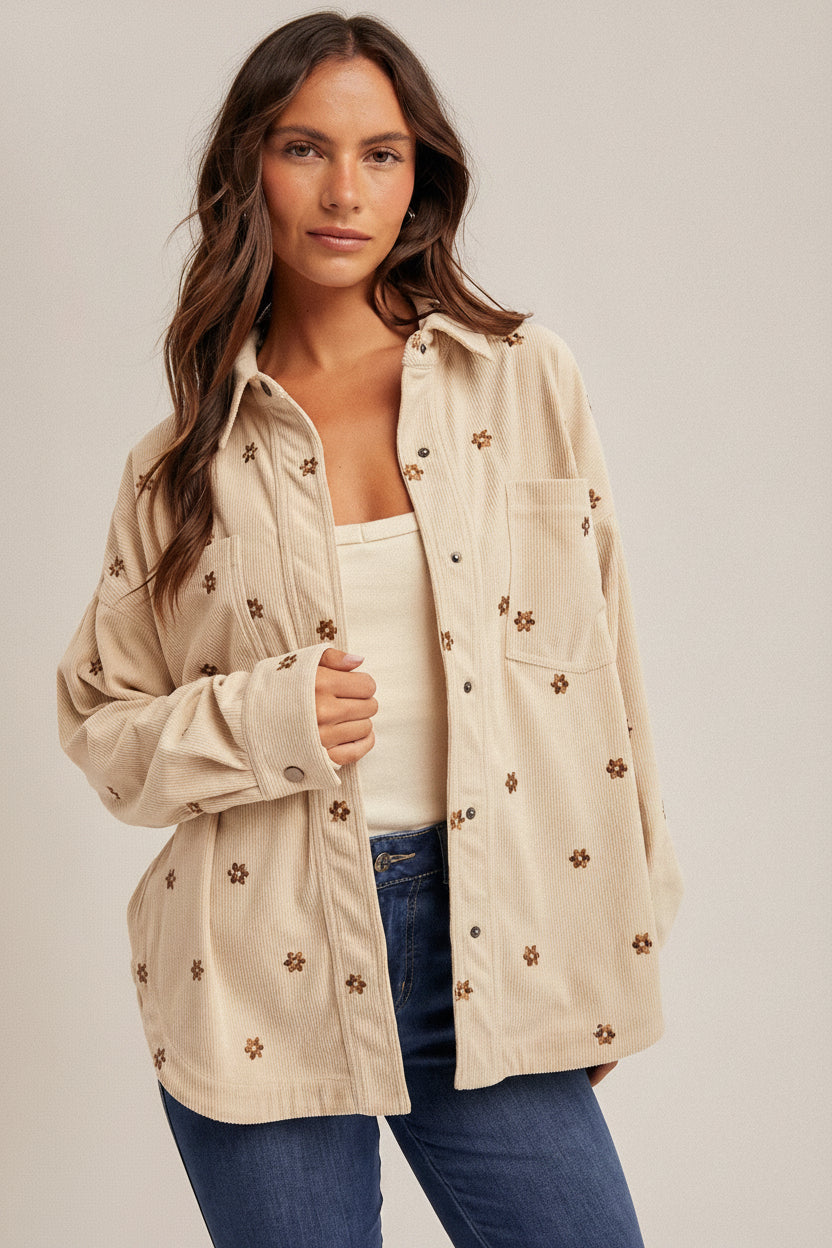 Woman wearing an ecru shirt jacket with brown metallic embroidery floral patterns, brown hat, and blue jeans on a plain background