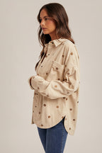 Woman wearing an ecru shirt jacket with brown metallic embroidery floral patterns, brown hat, and blue jeans on a plain background