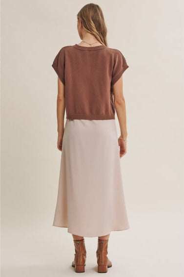 Back view of a woman wearing a mocha dolman sleeve cable-knit cropped top and a champagne A-line satin slip midi dress, styled with brown ankle boots against a plain beige studio background.