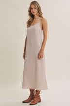A woman wearing a champagne A-line satin slip midi dress with spaghetti straps, styled with brown ankle boots against a plain beige studio background.