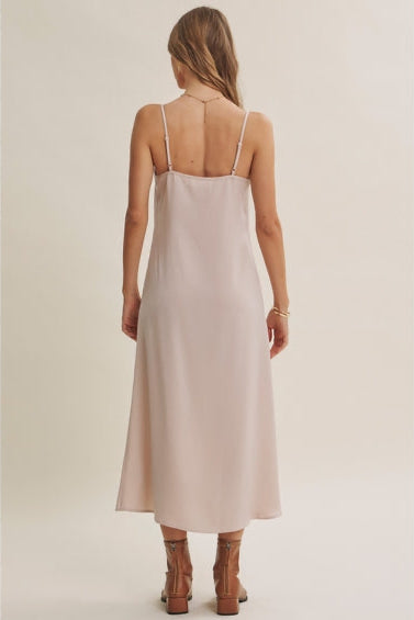 Back view of a woman wearing a champagne A-line satin slip midi dress with spaghetti straps, styled with brown ankle boots against a plain beige studio background.