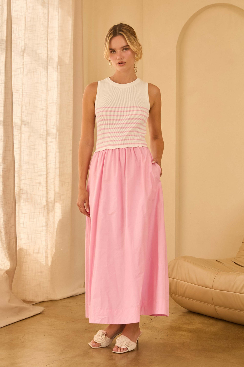 Woman wearing a maxi dress with pink and white striped top and a long pink skirt standing against beige walls 