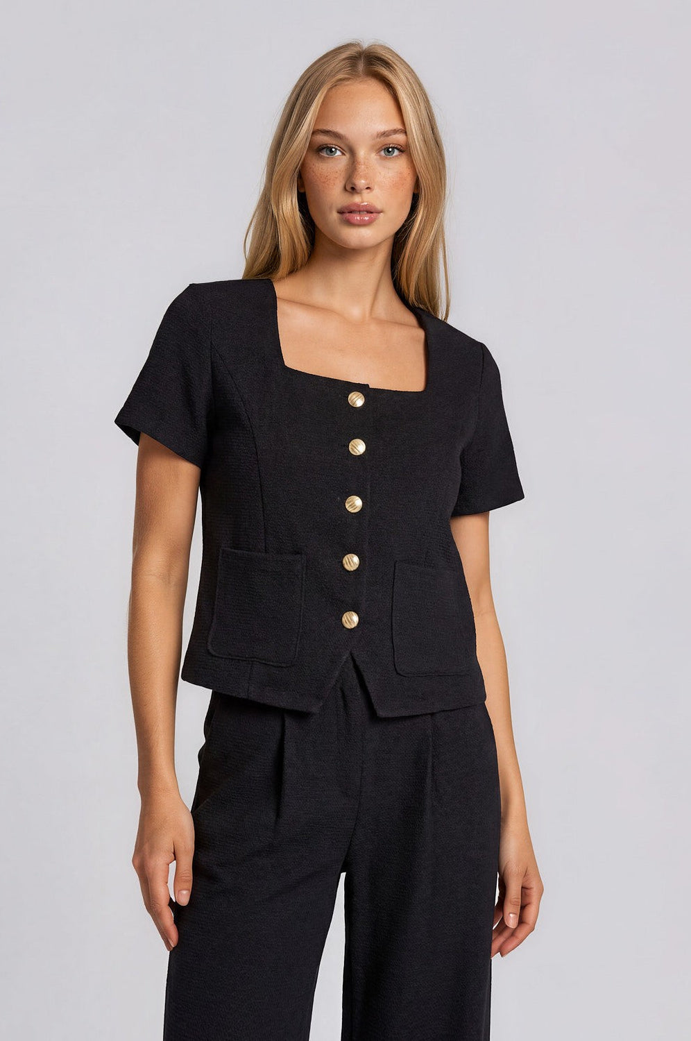 A woman wearing a black short-sleeve  top with a square neckline, gold front button detail, and front pockets, paired with black trousers, against a light gray studio background.