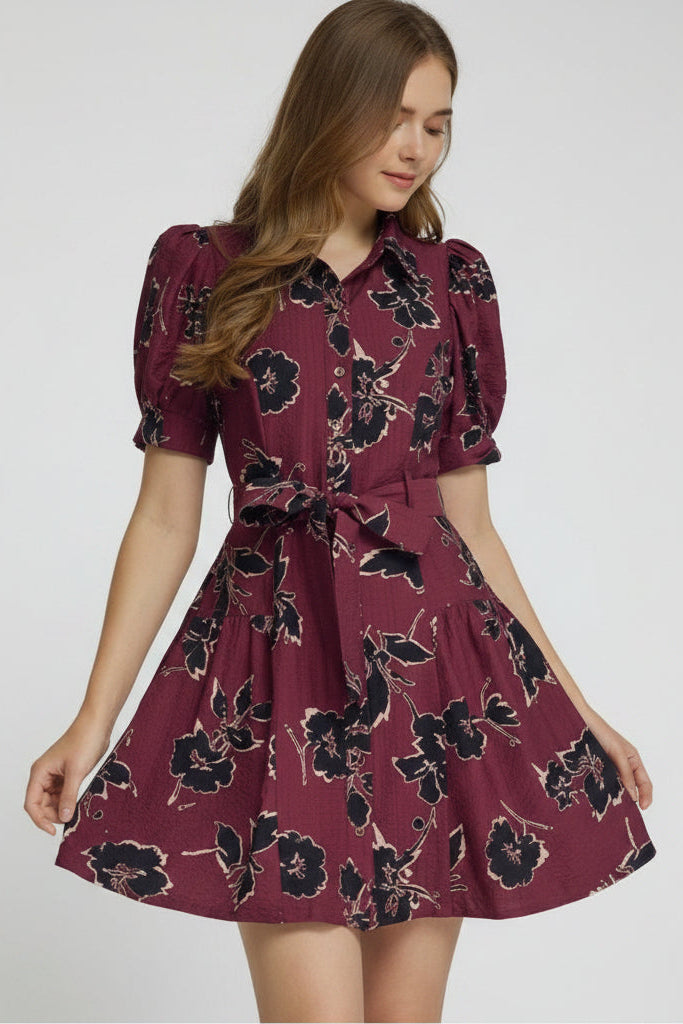 A woman modeling a short puff-sleeve wine dress with a black floral print, featuring a button-front bodice and a matching tie belt at the waist, set against a plain light gray studio background.