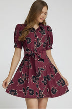 A woman modeling a short puff-sleeve wine dress with a black floral print, featuring a button-front bodice and a matching tie belt at the waist, set against a plain light gray studio background.