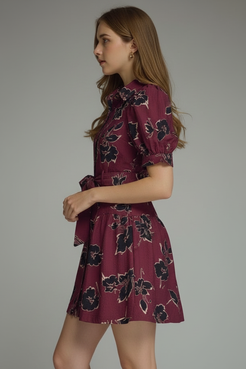 A woman modeling a short puff-sleeve wine dress with a black floral print, featuring a button-front bodice and a matching tie belt at the waist, set against a plain light gray studio background.