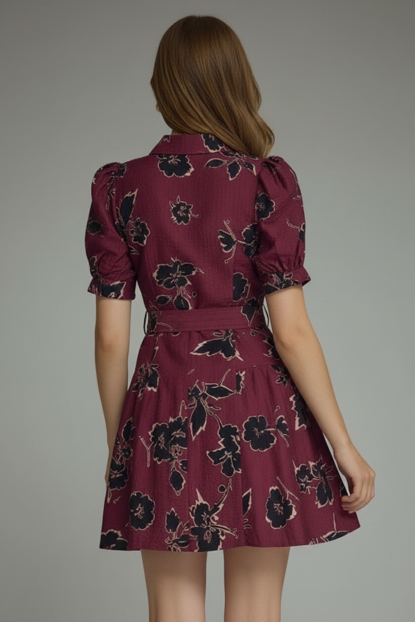 Back view of a woman modeling a short puff-sleeve wine dress with a black floral print, featuring a button-front bodice and a matching tie belt at the waist, set against a plain light gray studio background.