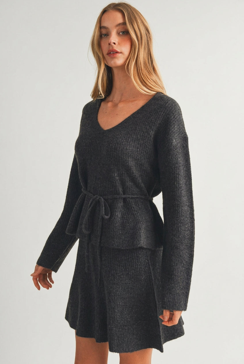 A woman modeling a charcoal gray ribbed knit top with long sleeves, a V-neckline, and a matching tie belt at the waist, set against a plain beige studio background.