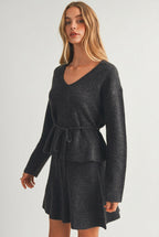 A woman modeling a charcoal gray ribbed knit top with long sleeves, a V-neckline, and a matching tie belt at the waist, set against a plain beige studio background.
