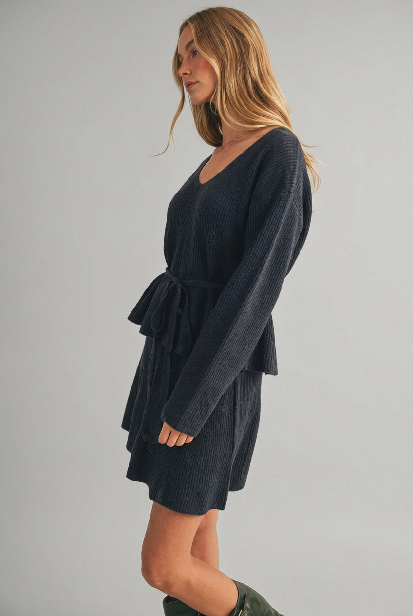 A woman modeling a charcoal gray ribbed knit top with long sleeves, a V-neckline, and a matching tie belt at the waist, set against a plain beige studio background.
