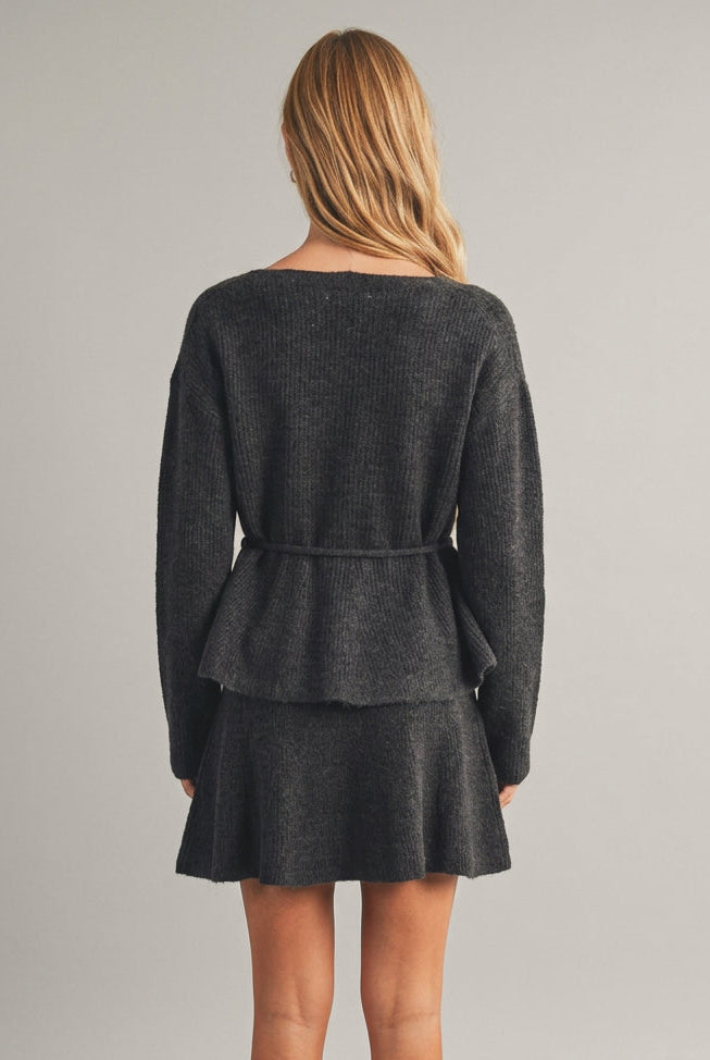 Back view of a woman modeling a charcoal gray ribbed knit top with long sleeves, a V-neckline, and a matching tie belt at the waist, set against a plain beige studio background.