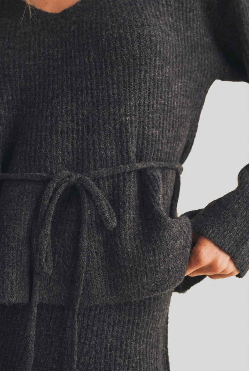 A woman modeling a charcoal gray ribbed knit top with long sleeves, a V-neckline, and a matching tie belt at the waist, set against a plain beige studio background.