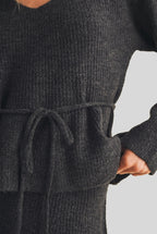 A woman modeling a charcoal gray ribbed knit top with long sleeves, a V-neckline, and a matching tie belt at the waist, set against a plain beige studio background.