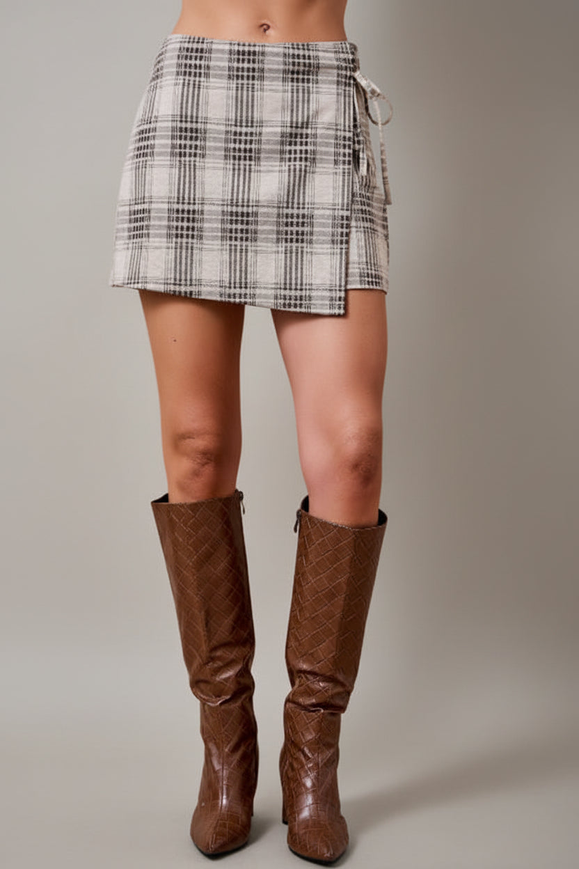 A woman wearing an oatmeal brown and cream plaid wrap skort with a side tie, paired with brown knee-high textured boots, set against a solid tan studio background.