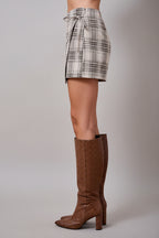 A woman wearing an oatmeal brown and cream plaid wrap skort with a side tie, paired with brown knee-high textured boots, set against a solid tan studio background.