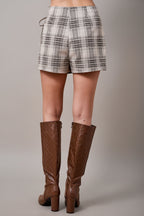 Back view of a woman wearing an oatmeal brown and cream plaid wrap skort with a side tie, paired with brown knee-high textured boots, set against a solid tan studio background.