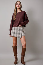 A woman wearing an oatmeal brown and cream plaid wrap skort with a side tie, paired with brown sweater and brown knee-high textured boots, set against a solid tan studio background.