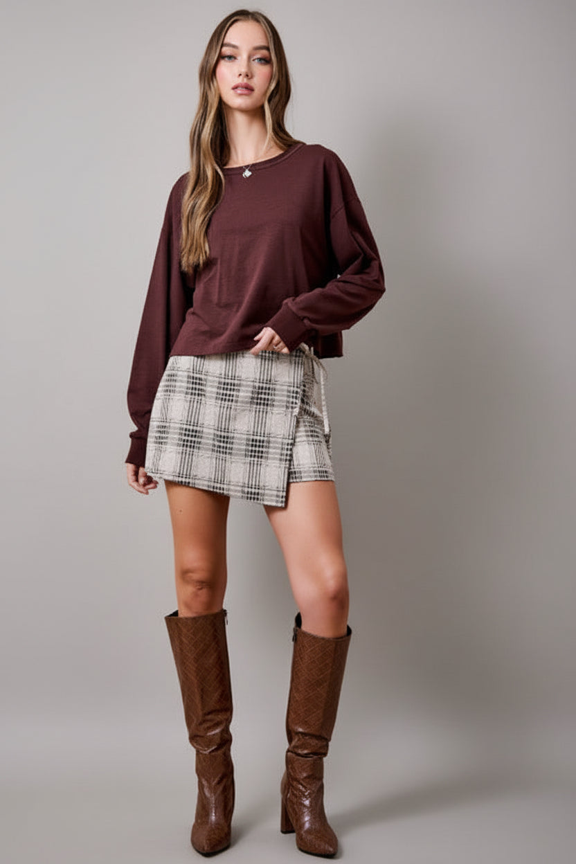 A woman wearing an oatmeal brown and cream plaid wrap skort with a side tie, paired with brown sweater and brown knee-high textured boots, set against a solid tan studio background.